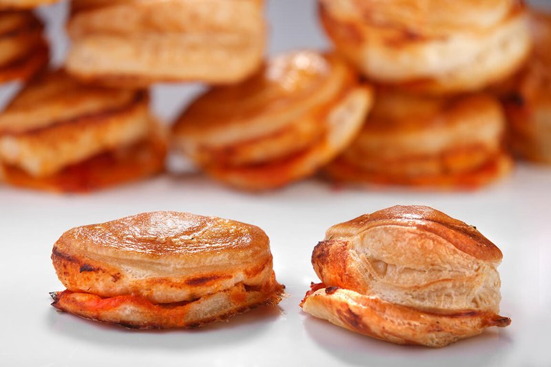 Golden-brown puff pastry pizzette with a crispy, flaky texture and a rich tomato filling, displayed on a white surface with more pizzette in the background. A delicious, bite-sized Italian snack.